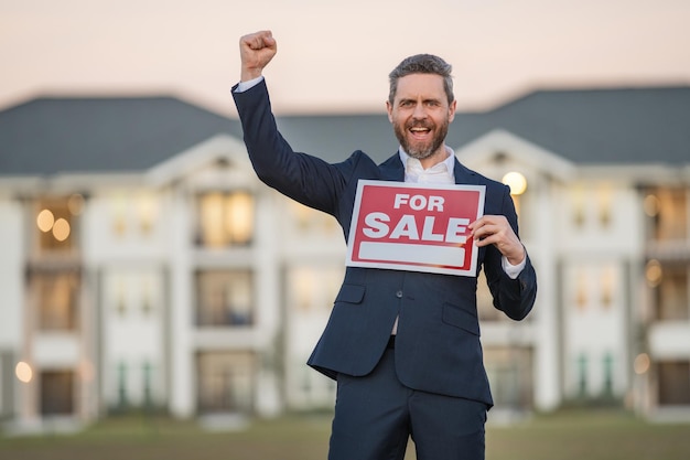 Erfolgreicher Immobilienmakler im Anzug mit Verkaufsschild Immobilienmakler vor Neubauhaus real e