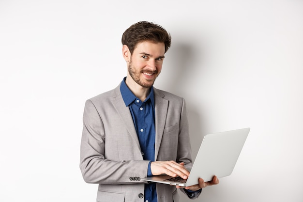 Erfolgreicher lächelnder Geschäftsmann, der auf Laptop arbeitet und glücklich auf Kamera schaut, im grauen Anzug auf weißem Hintergrund stehend.