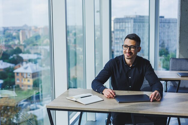 Erfolgreicher männlicher Geschäftsmann mit Brille und schwarzem Hemd sitzt an einem Schreibtisch und arbeitet über einen Laptop in einem modernen Büro an einem Projekt und blickt auf den Bildschirm