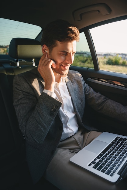 Erfolgreicher Mann, der Anzug und Ohrhörer trägt, der am Laptop arbeitet, während zurück im Geschäftsklassenauto sitzt