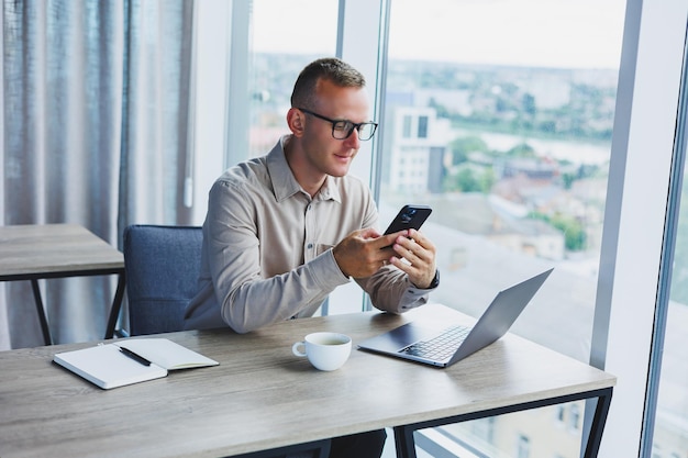 Erfolgreicher Mann, der auf dem Smartphone spricht und im Netbook surft, während er im Büro am Tisch im Internet surft