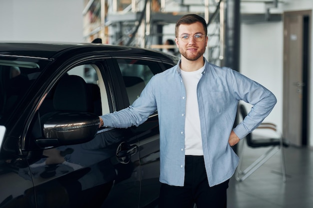 Erfolgreicher Mann mit Brille, der drinnen in der Nähe eines brandneuen Autos steht