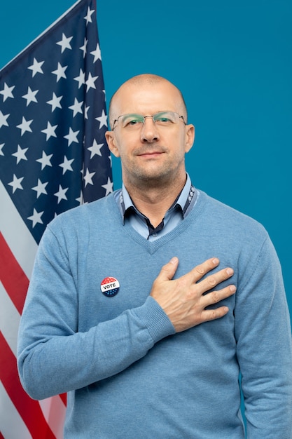 Erfolgreicher reifer Geschäftsmann, der rechte Hand auf Brust hält, während er vor Kamera gegen amerikanische Flagge und Blau steht