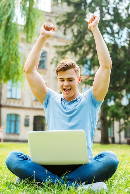 Erfolgreicher Schüler. Glücklicher junger Mann, der auf den Laptop schaut und seine Arme hebt, während er auf dem Gras sitzt