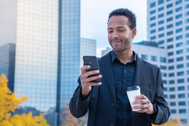 Erfolgreicher schwarzer Geschäftsmann im Anzug lächelt mit Smartphone und Kaffeetasse im Finanzviertel
