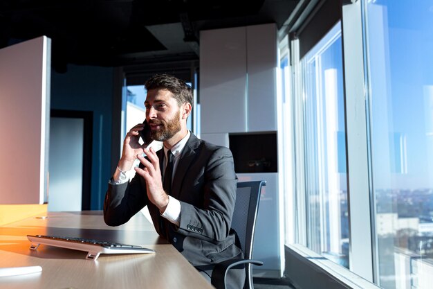 Erfolgreicher und glücklicher Geschäftsmann, der im modernen Büro am Computer arbeitet, Mann, der Spaß am Telefonieren hat