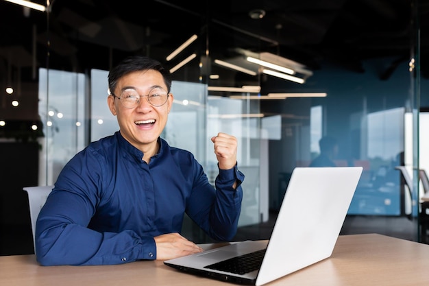 Erfolgreicher und glücklicher junger asiatischer Geschäftsmann, der im Büro am Laptop sitzt, freut sich über einen Gewinn a