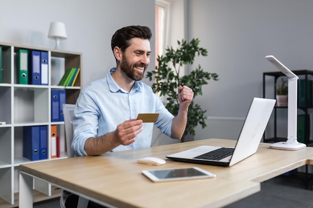 Erfolgreiches Online-Shopping Erfolgreiches Geschäft Zufriedener junger Mann, der eine Kreditkarte hält, die seine Hand zeigt Ja Sitzt an einem Schreibtisch im Büro am Computer