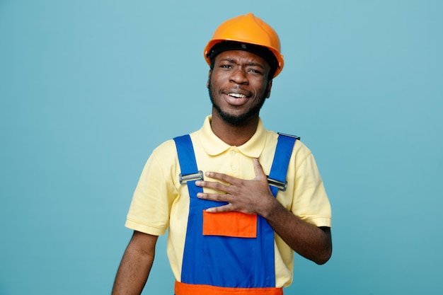 Erfreut, die Hand auf das Herz des jungen afroamerikanischen Baumeisters in Uniform zu legen, der auf blauem Hintergrund isoliert ist