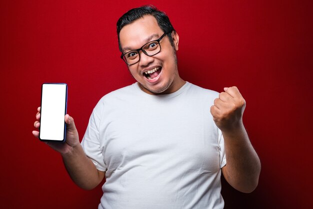 Erfreut glücklicher junger asiatischer Kerl im weißen T-Shirt, der in die Kamera lächelt, während er das Smartphone hält, das auf den Handybildschirm über rotem Hintergrund zeigt