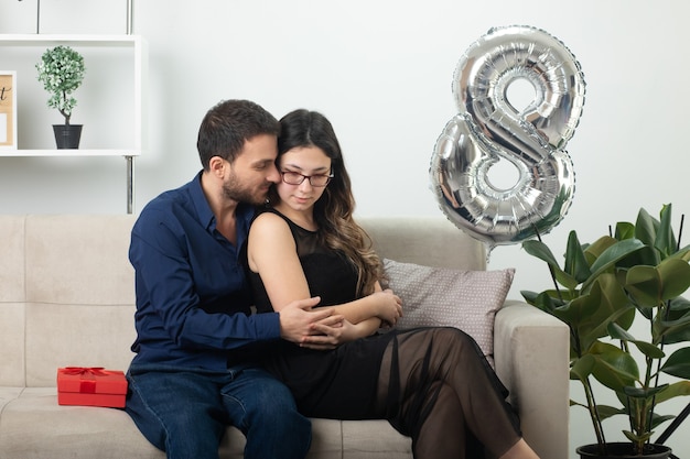 Erfreut, gutaussehender Mann, der am internationalen Frauentag im März eine hübsche junge Frau mit Brille auf der Couch im Wohnzimmer umarmt und anschaut