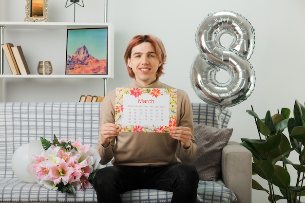 Erfreut hübscher Kerl am glücklichen Frauentag, der den Kalender auf dem Sofa im Wohnzimmer hält