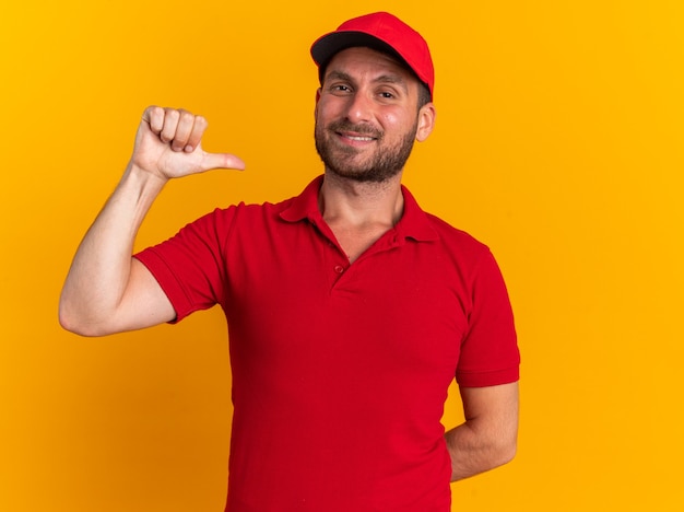 Foto erfreut junger kaukasischer liefermann in roter uniform und mütze, der die hand hinter dem rücken hält und auf die kamera schaut, die auf sich selbst zeigt, isoliert auf oranger wand