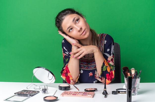 Erfreut junges brünettes Mädchen, das am Tisch mit Make-up-Tools sitzt, die Hand auf ihr Gesicht legt und nach oben schaut