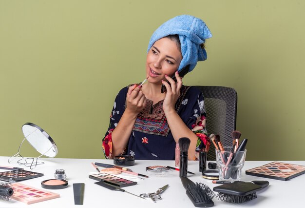 Erfreut junges brünettes Mädchen mit eingewickelten Haaren in Handtuch, das am Tisch mit Make-up-Tools sitzt und am Telefon spricht und Lipgloss auf olivgrüner Wand mit Kopienraum auftragt