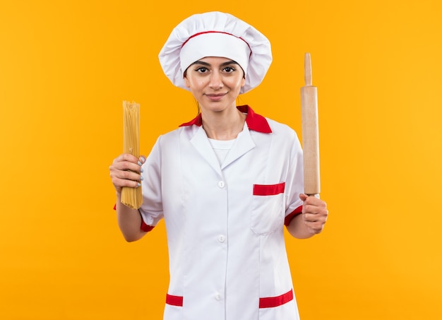 Erfreut junges schönes Mädchen in Kochuniform mit Spaghetti mit Nudelholz isoliert auf oranger Wand
