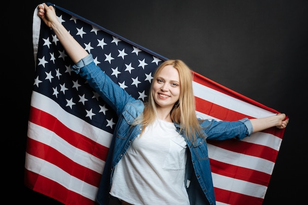 Erfreute amüsierte junge Frau, die Spaß hat und Freude ausdrückt, während sie Nationalflagge hält und isoliert in der schwarzen Wand steht