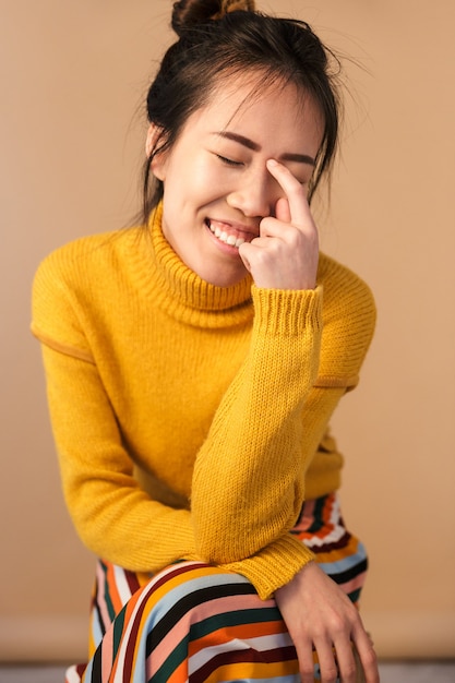 erfreute asiatische Frau, die einen Pullover trägt und lächelt, während sie im Studio sitzt, isoliert über beige Wand