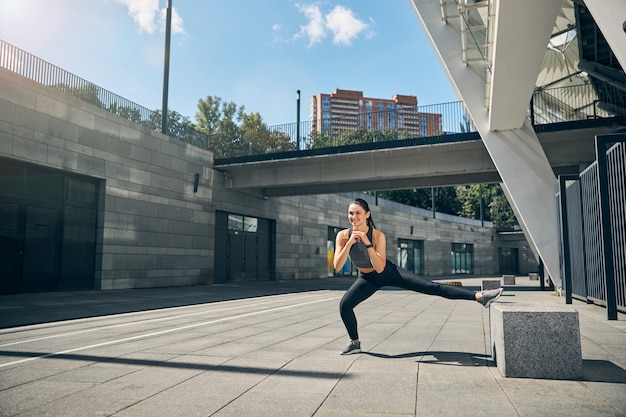 Erfreute brünette Frau, die ein Lächeln auf ihrem Gesicht behält, während sie alleine im Freien Sport treibt