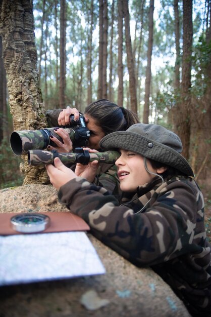 Foto erfreute familie beim fotografieren im wald. mutter und sohn mit modernen kameras, die auf dem boden liegen und aus stein gucken. elternschaft, familie, freizeitkonzept