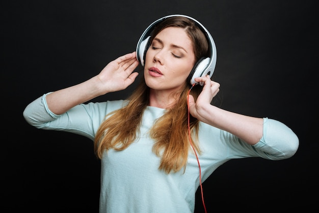 Erfreute junge friedliche Frau, die Friedlichkeit ausdrückt und Kopfhörer benutzt, während sie die Musik hört und isoliert in der schwarzen Wand steht