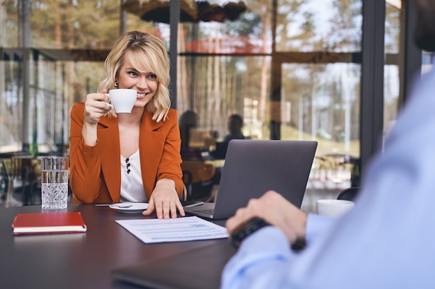 Erfreute junge Unternehmerin mit einer Kaffeetasse, die vor ihrem Geschäftspartner sitzt