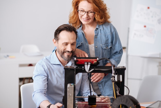 Erfreute professionelle 3D-Designer, die lächeln und den 3D-Drucker betrachten, während sie zusammenarbeiten
