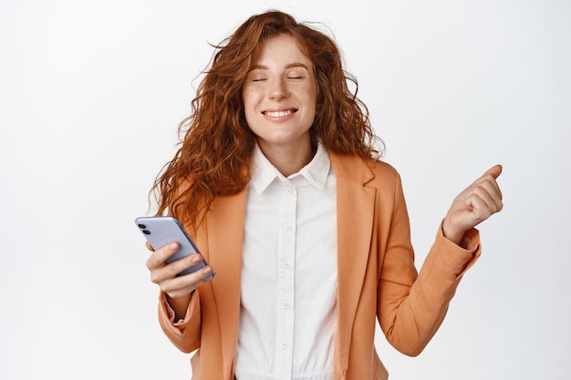 Erfreute Verkäuferin, die ihr Telefon hält und triumphierend lächelnd zufrieden im Anzug vor weißem Hintergrund steht