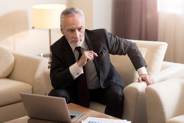 Erfreuter alter bärtiger Geschäftsmann, der im Hotel vor dem Laptop sitzt und voller Ideen ist, während er Interesse ausdrückt