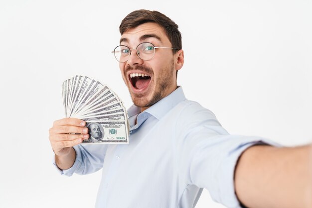 erfreuter Mann mit Brille, der Dollar hält und ein Selfie-Foto macht, das über weißer Wand isoliert ist?