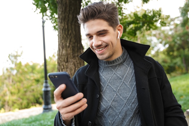 erfreuter optimistischer, gutaussehender junger Mann in Freizeitkleidung, der draußen im grünen Park spazieren geht und Handy-Musik mit Kopfhörern hört