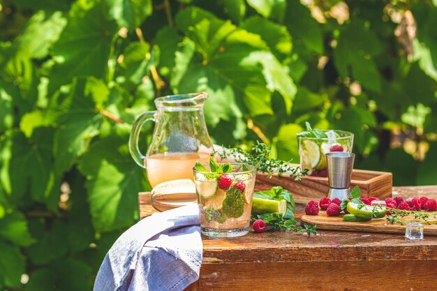 Erfrischender Minz-Cocktail-Mojito mit Rum-Limette und Himbeer-Kaltgetränk oder Getränk auf einem rustikalen Holztisch im Freien. Sonniger Tag mit grünen Blättern im Hintergrund