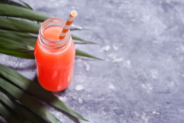 Erfrischender tropischer Sommercocktail in einer Flasche mit Strohhalm auf einem grauen Betontisch unter dem Palmblatt.