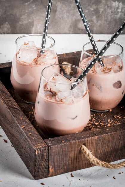 Erfrischungsgetränke im Sommer. Gekühlter gefrorener Schokoladenkakao. Mit einer Kugel Schokoladeneis, Schokoladenpulver und Eis. In Gläsern mit Röhrchen zum Trinken. Holztablett der weißen konkreten Tabelle.