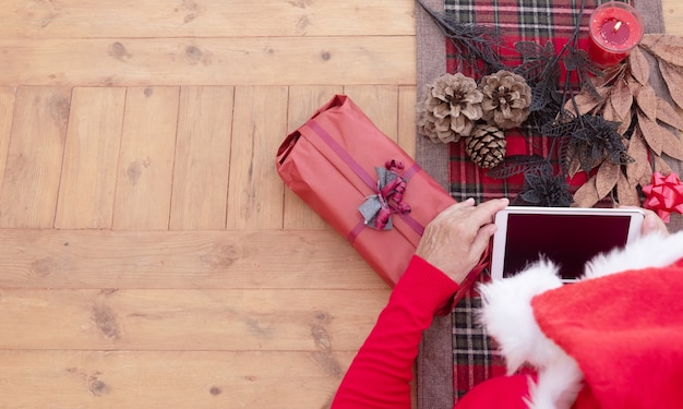 Erhöhte Ansicht der Hände einer älteren Frau, die eine Weihnachtsmütze mit Technologie mit einem kleinen Tablet trägt. Holztisch dekoriert mit Weihnachtsaccessoires