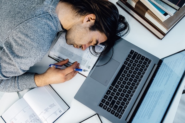 Erhöhte Ansicht des müden Geschäftsmannes, der schläft, während er die Ausgaben am Schreibtisch im Büro berechnet. Junge kaukasische männliche Angestellte machen ein Nickerchen im Büro auf einem Tisch mit Tablet- und Laptop-Computer.