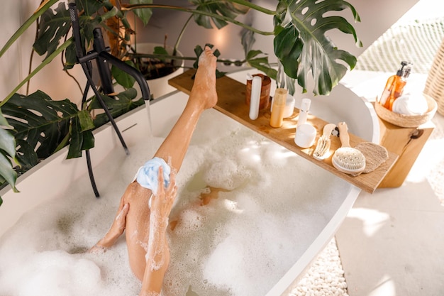 Erhöhte Ansicht einer jungen Frau, die einen Schwamm benutzt, während sie sich in einer mit Blasen gefüllten Badewanne entspannt
