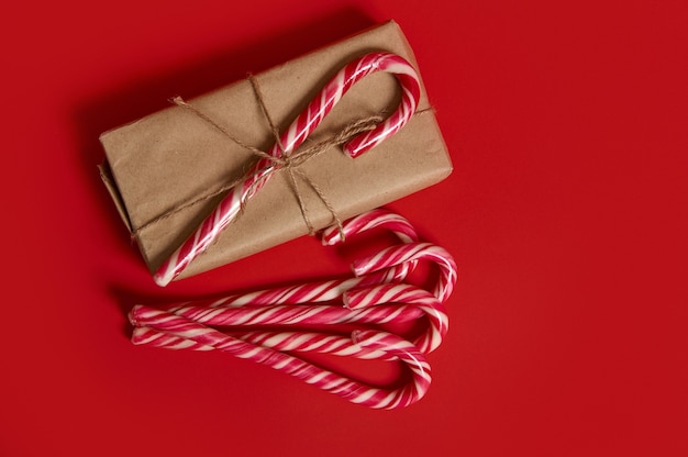 Erhöhte Ansicht von traditionellen Weihnachtsartikeln. Gestreifte Zuckerstange auf einem Geschenk verpackt in Kraftpapier und Lutscher auf rotem Hintergrund mit Kopienraum für Neujahrswerbung