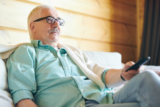 Erholsamer älterer Mann in Brille und Freizeitkleidung mit Fernbedienung beim Fernsehen in seinem Landhaus