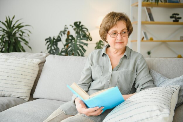 Erholsames Lesen Porträt einer nachdenklichen alten Frau, die Lieblingsliteratur in einem gemütlichen Zuhause liest. Sie liegt auf Kissen auf einem bequemen Sofa. Die Beine sind mit einer weichen Decke bedeckt