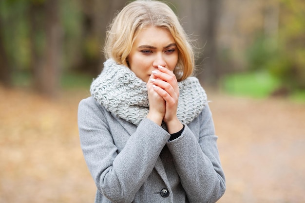Erkältung und Grippe. Junge Frau in einem grauen Mantel, die im Herbstpark spaziert und gefrorene Hand wärmt.