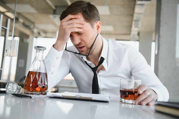 Ermüdender junger Geschäftsmann, der am Arbeitsplatz sitzt und Whiskeyflasche hält, Alkoholkonkurskonzept trinkt