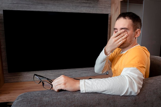 Ermüdung des Sehens Ein Zuschauer in einem gelben Hemd auf dem Hintergrund des Fernsehmanns fühlt sich müde, wenn er die Augen berührt, wenn er zu Hause fernsieht