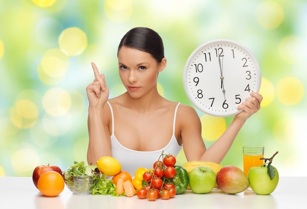 Ernährung, gesunde Ernährung, Zeit, Abnehmen und Menschen Konzept - Frau mit Essen auf dem Tisch und große Uhr sitzt am Tisch über grünen Sommerlichtern Hintergrund