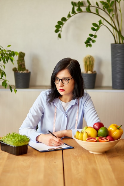 Ernährungsberaterin, Diätassistentin, die einen Ernährungsplan schreibt, mit gesundem Gemüse und Obst, Gesundheits- und Ernährungskonzept. Ernährungsberaterin mit Früchten, die an ihrem Schreibtisch arbeiten.