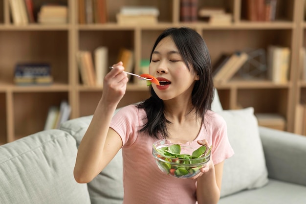 Ernährungskonzept Junge koreanische Dame, die gesundes Essen isst und eine Schüssel mit frischem Salat hält, die zu Hause auf dem Sofa sitzt, freier Platz