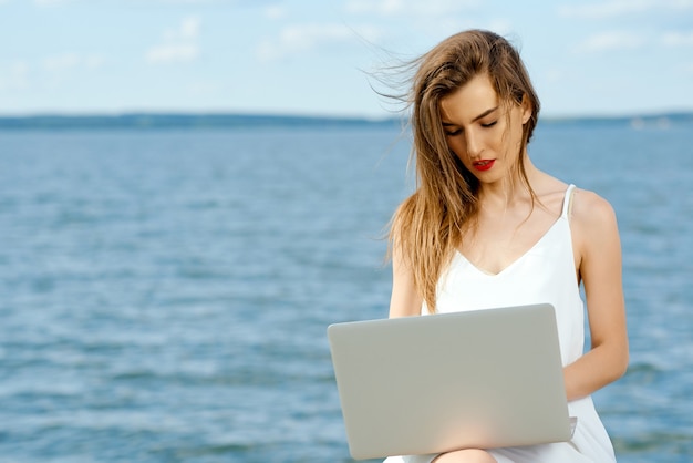 Ernste Frau mit Laptop im weißen Kleid sitzt vor dem Hintergrund des Wassers. Foto in hoher Qualität