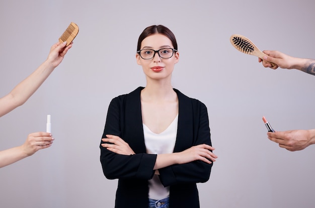 Ernste Geschäftsfrau steht auf einer grauen Rückseite in einer schwarzen Jacke, in einem weißen T-Shirt und in einer Computerbrille. Ihre Arme verschränkt. Die falschen Hände geben ihr zwei Kämme und zwei Lippenstifte.