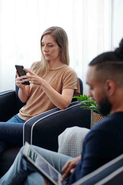 Ernste hübsche junge Frau, die im Sessel neben ihrem Ehemann sitzt und Textnachrichten auf dem Smartphone beantwortet oder Artikel liest