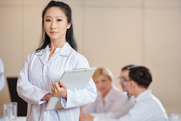 Ernste junge chinesische Wissenschaftlerin mit Zwischenablage, die im Konferenzraum steht und in die Kamera schaut